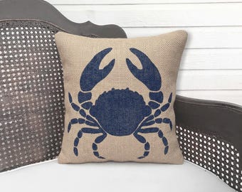 May include: A square throw pillow featuring a textured tan fabric base with a prominent dark blue graphic of a crab centered on the front. The crab design displays two large claws pointing upward and multiple legs extending from the central body.