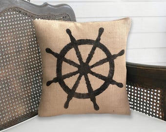May include: A square throw pillow made of tan, textured burlap fabric. The center of the pillow features a large, bold, dark brown graphic print of a ship's steering wheel with eight spokes, centered symmetrically on the front surface.