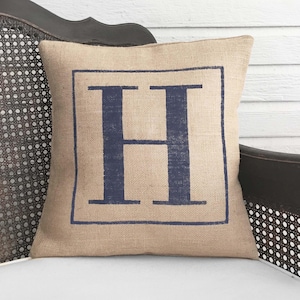 May include: A burlap pillow with a navy blue letter "H" printed in a square on the front.