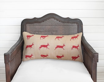 May include: A rectangular lumbar pillow featuring a tan, textured fabric base with a repeating pattern of twelve red deer silhouettes printed in a grid layout. The deer are depicted in various running poses across the surface of the pillow.