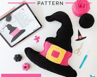 May include: A black crochet witch hat pillow with a pink and yellow band. The pillow is on a white surface with other Halloween-themed items, including a pink yarn ball, a black yarn ball, a ghost cutout, a small pumpkin, a small black pumpkin, a pair of scissors, a crochet hook, and a witch hat cutout. The image also includes the text "Witch Hat Crochet Pillow Pattern" and "PDF Download".
