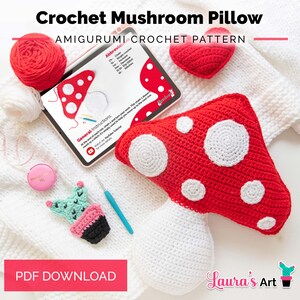 Puede incluir: Un cojín de ganchillo con forma de seta, con sombrero rojo de puntos blancos y tallo blanco, una tableta con un patrón, un corazón rojo, un cactus pequeño, ovillos de lana roja y blanca, una cinta métrica rosa y un ganchillo azul.