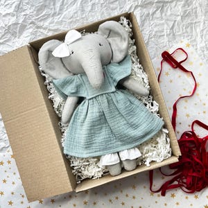 May include: A gray stuffed elephant doll wearing a light blue dress with white trim, a white bow, and a red heart. The doll is in a cardboard box filled with white paper shreds.