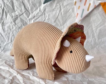 May include: A plush toy triceratops in a beige corduroy fabric with a colorful patterned frill. The toy has a white horn and black eyes.