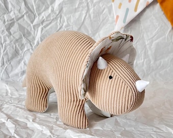 May include: A plush brown corduroy triceratops toy with a white floral print on its frill. The toy has white horns and a black eye.