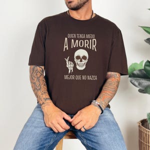 May include: A brown t-shirt with the text "QUIEN TENGA MIEDO A MORIR MEJOR QUE NO NAZCA" in white, along with a skull and hand bone graphic. The shirt is a short-sleeved crew neck.