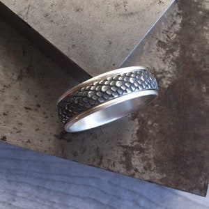 May include: A silver ring with a textured black band. The band has a pattern of small, raised squares.