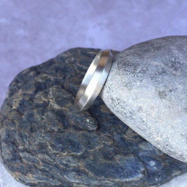 Peut inclure: Une bague en argent avec un bord plat et biseaut&eacute;. La bague repose sur une roche grise.