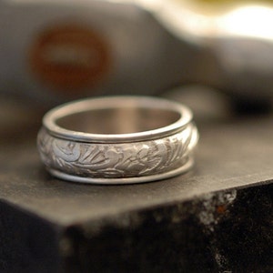 May include: A silver wedding band with an intricate floral design engraved around the band.