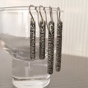 May include: Four silver dangle earrings with a floral pattern. The earrings are hanging from a glass surface.