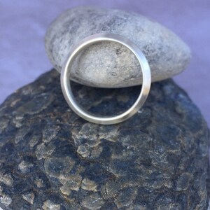Peut inclure: Une bague simple en argent, avec une finition lisse et polie. La bague est pr&eacute;sent&eacute;e sur une pile de rochers gris.