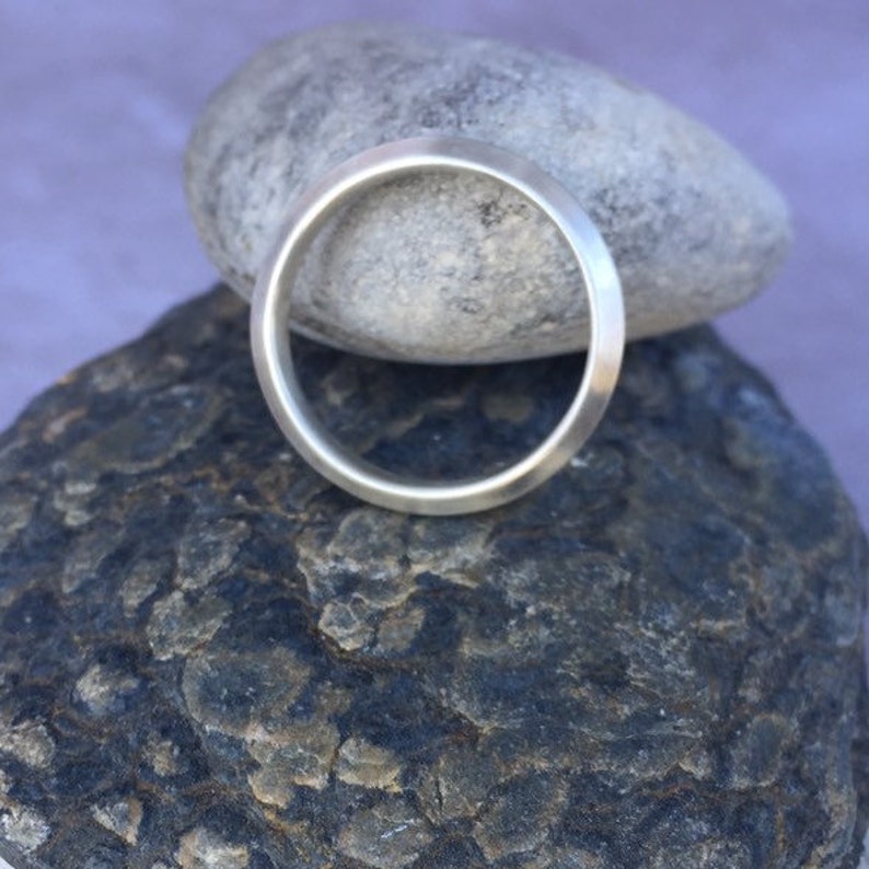 Peut inclure: Une bague simple en argent, avec une finition lisse et polie. La bague est pr&eacute;sent&eacute;e sur une pile de rochers gris.