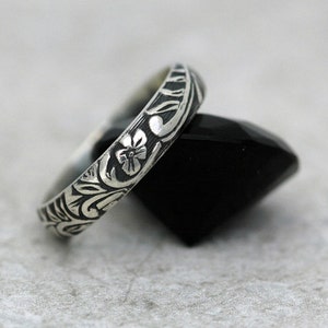 May include: A silver band ring with a floral design. The ring is engraved with a delicate pattern of leaves and flowers.