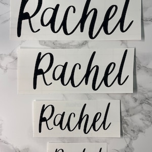 May include: Four white sheets of paper with the name "Rachel" written in black cursive lettering. The sheets are arranged in a vertical stack, with the largest sheet at the top and the smallest sheet at the bottom.