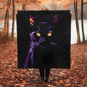 Peut inclure: Couverture noire avec un motif de chat stylisé. Le chat a des yeux jaunes, des oreilles et des moustaches violettes, sur fond noir. La couverture est tenue devant une personne dans un cadre forestier.