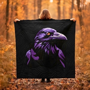 Op de afbeelding: Een zwarte quilt met een gestileerd paars ravenmotief. De raaf heeft oranje ogen en staat tegen een zwarte achtergrond met een gewatteerd patroon. De afbeelding is gemaakt in een herfstbos.