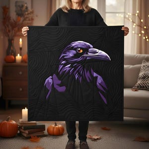 Op de afbeelding: Een zwarte quilt met een paars ravenontwerp en oranje ogen. De raaf is de centrale afbeelding op de quilt, die een gestructureerd, golvend patroon heeft. De quilt wordt vastgehouden door een persoon in een kamer met pompoenen en kaarsen.