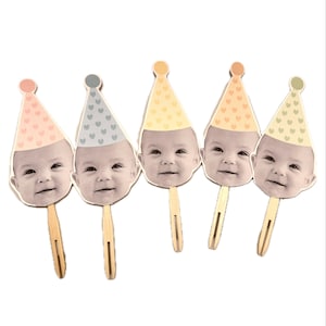 May include: Five cupcake toppers featuring baby faces with different colored party hats. The hats are decorated with small white hearts. The toppers are made of cardstock and have wooden sticks.
