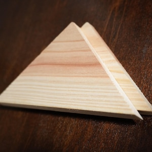 May include: Two wooden triangles stacked on top of each other, with the grain of the wood visible. The triangles are light brown in color and are sitting on a dark brown surface.