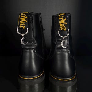 Może przedstawiać: Para czarnych skórzanych butów z żółtymi przeszyciami i grubą podeszwą. Każdy but ma srebrny urok z motywem półksiężyca wiszącym na pętli, która ma żółty napis "AirWair".