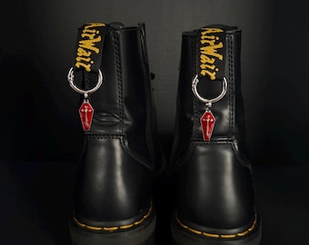 Könnte beinhalten: Paar schwarze Lederstiefel mit gelben Nähten und dicken Sohlen. Jeder Stiefel hat einen roten Sarganhänger mit einem silbernen Ring und einem gelb-schwarzen "AirWair"-Etikett.