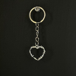 May include: A silver-colored keychain with a heart-shaped charm. The heart is crafted from wire, with a barbed wire design. The keychain includes a chain and a circular key ring. The background is black.