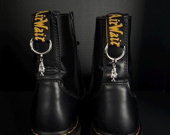 Könnte beinhalten: Paar schwarze Leder-Plateau-Stiefel mit gelben Nähten und einem gelb-schwarzen Zugband mit der Aufschrift "AirWair". Jeder Stiefel hat einen silbernen Anhänger, der an einem Metallring hängt.