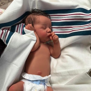 May include: A newborn baby doll with dark hair and rosy cheeks, wearing a diaper, lies swaddled in a white blanket with teal and burgundy stripes. The baby is resting with its eyes closed, and its hand is near its mouth.