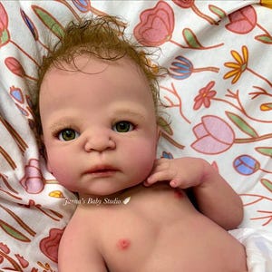 Może przedstawiać: Realistyczna lalka typu reborn o jasnej karnacji, zielonych oczach i rzadkich, cienkich brązowych włosach. Lalka ma neutralny wyraz twarzy, widoczne namalowane brwi i ma na sobie białą pieluchę. Na klatce piersiowej widoczny napis Janna's Baby Studio.