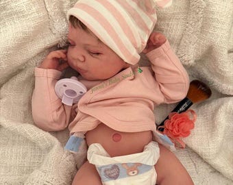 May include: A realistic reborn doll with brown hair wearing a pink and white striped knotted hat and a long-sleeved light pink wrap shirt. The doll has a white pacifier, a diaper with a rabbit graphic, and a small red belly button mark.