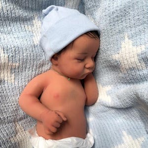 May include: A realistic baby doll with a light blue beanie and a diaper. The doll has dark hair and is lying on a blue blanket. The baby's skin tone is tan, and it appears to be sleeping.
