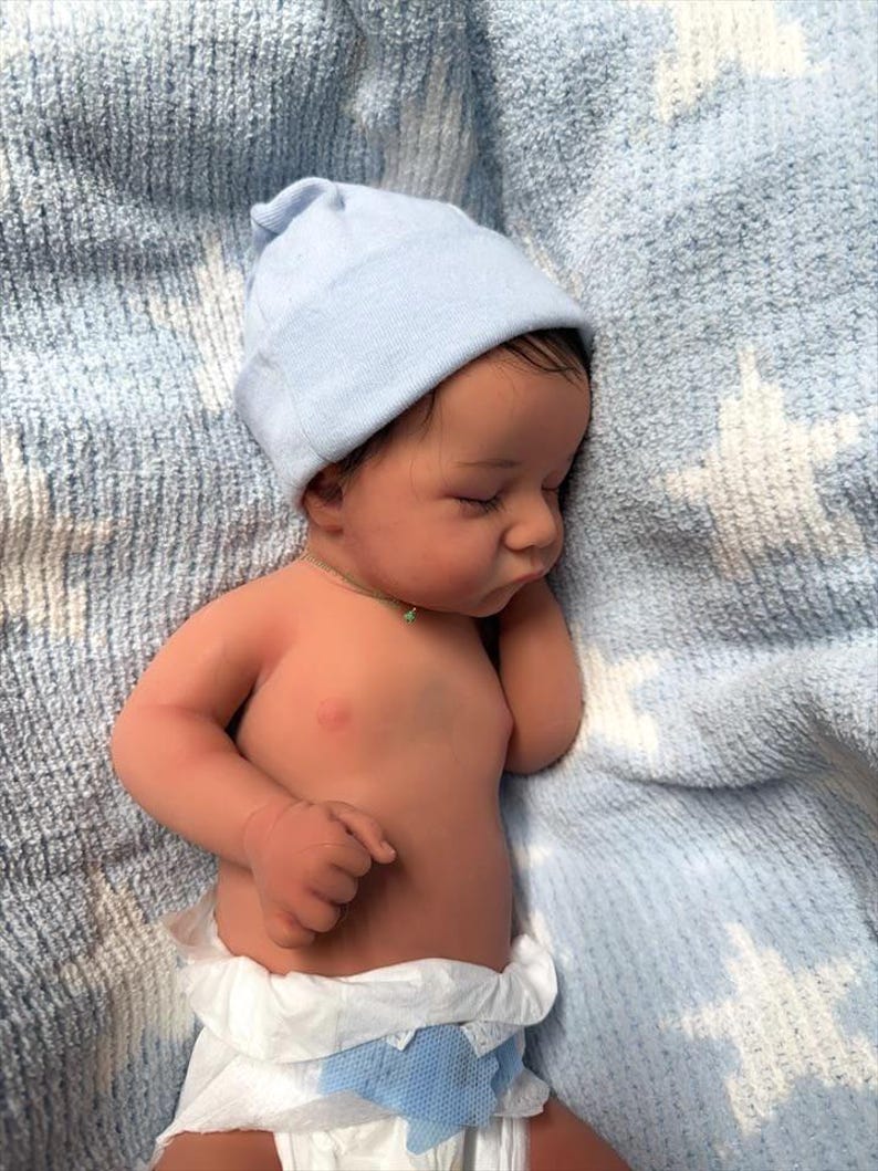 May include: A realistic baby doll with a light blue beanie and a diaper. The doll has dark hair and is lying on a blue blanket. The baby's skin tone is tan, and it appears to be sleeping.