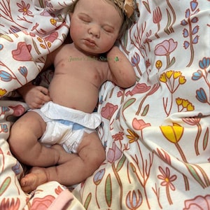 May include: A realistic baby doll with closed eyes, wearing a nappy, and a floral headband. The doll is nestled in a blanket with a colourful floral pattern. The doll's skin has a lifelike appearance.
