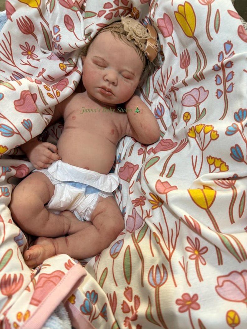 May include: A realistic baby doll with closed eyes, wearing a nappy, and a floral headband. The doll is nestled in a blanket with a colourful floral pattern. The doll's skin has a lifelike appearance.