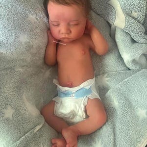 May include: A lifelike baby doll with closed eyes, wearing a diaper, and lying on a soft, light blue blanket. The doll has detailed skin tones and brown hair. The blanket has a star pattern.