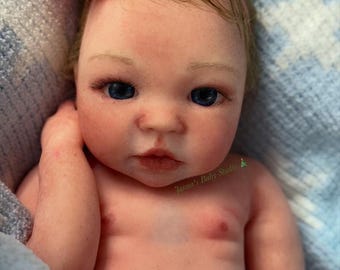 Puede incluir: Una muñeca bebé realista con ojos azules y tez color melocotón. La muñeca tiene una diadema de flores rosas. La muñeca está acostada sobre una manta azul. El texto "Janna's Baby Studio" es visible.
