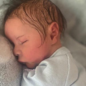 May include: A newborn baby with dark hair is sleeping peacefully, its face gently illuminated. The baby is wearing a light blue onesie and resting on a soft, gray blanket. The text "Janna's Baby Studio" is visible.