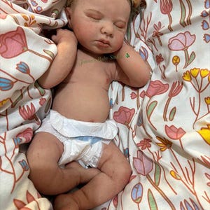 May include: A lifelike baby doll with closed eyes, wearing a nappy and a headband with a bow. The doll is lying on a floral patterned blanket with pink, yellow, and blue flowers. The text "Janna's Baby Studio" is visible.