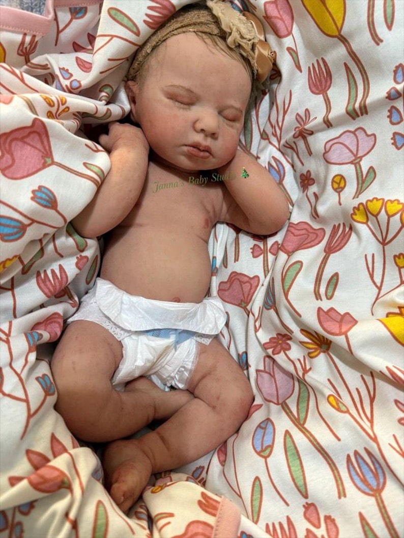 May include: A lifelike baby doll with closed eyes, wearing a nappy and a headband with a bow. The doll is lying on a floral patterned blanket with pink, yellow, and blue flowers. The text "Janna's Baby Studio" is visible.