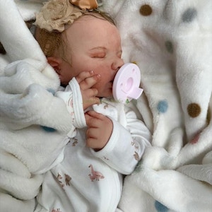 May include: A lifelike baby doll, dressed in a white onesie featuring pink flamingo and bear designs, and a matching bow. The doll is wrapped in a soft, white blanket with polka dots, and is using a pink dummy.