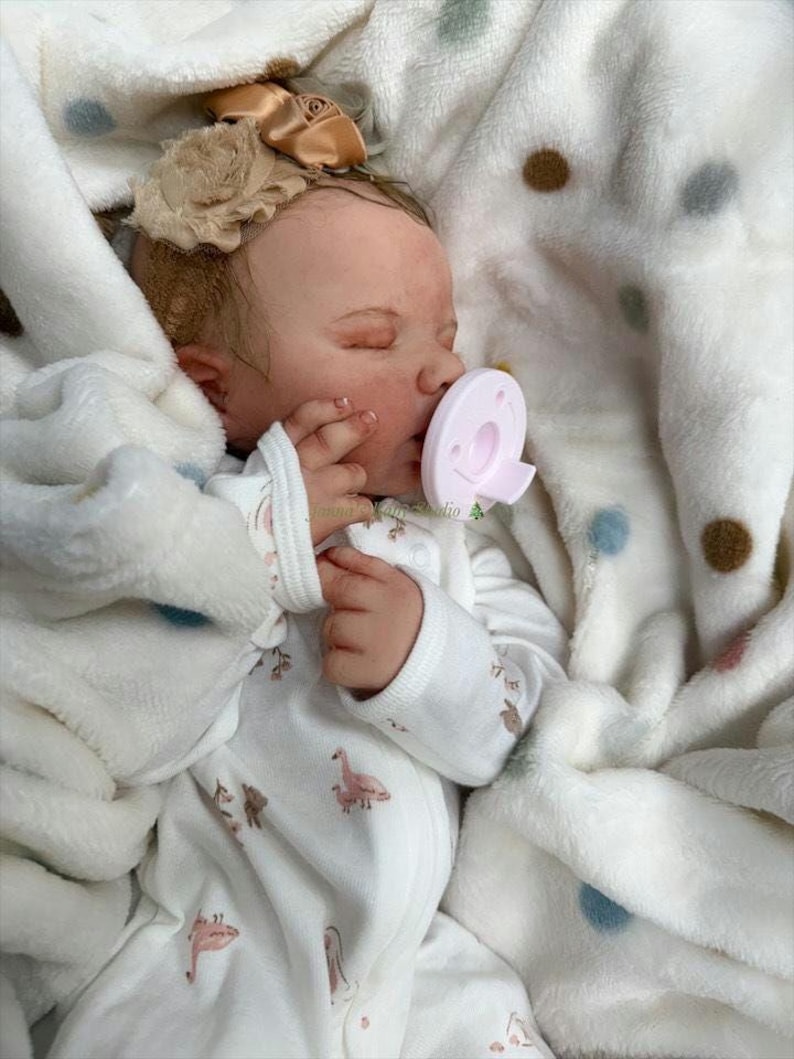 May include: A lifelike baby doll, dressed in a white onesie featuring pink flamingo and bear designs, and a matching bow. The doll is wrapped in a soft, white blanket with polka dots, and is using a pink dummy.