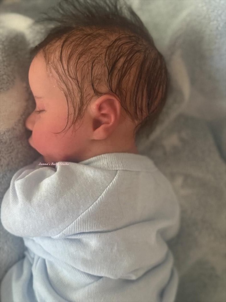 May include: A sleeping baby with dark hair and rosy cheeks, wearing a light blue onesie. The baby is lying on a soft, light blue blanket. The text "Janna's Baby Studio" is visible on the image.