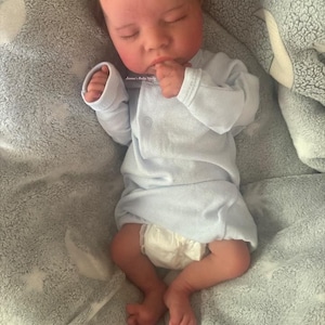 May include: A realistic baby doll is lying on a gray blanket. The doll is wearing a light blue onesie and a diaper. The baby has closed eyes and is resting its hand near its mouth. The blanket has a star pattern.