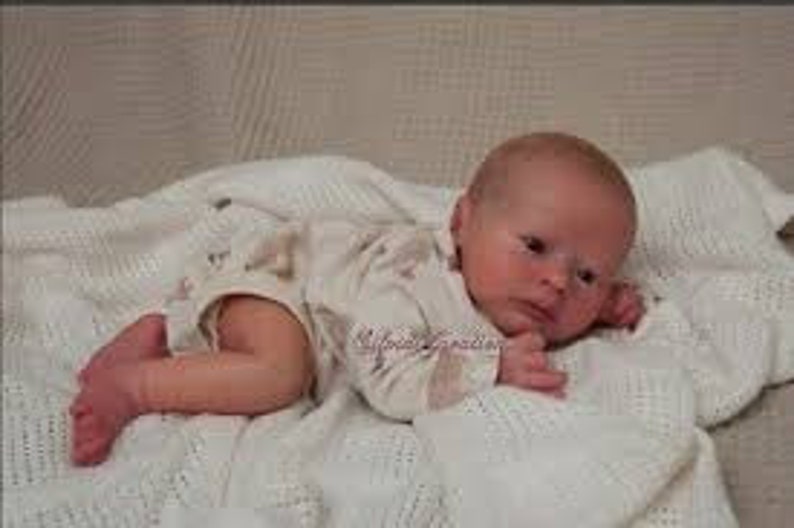 May include: A realistic baby doll with a light brown complexion, brown eyes, and a light brown wig. The doll is wearing a white and brown patterned onesie and is lying on a white blanket.