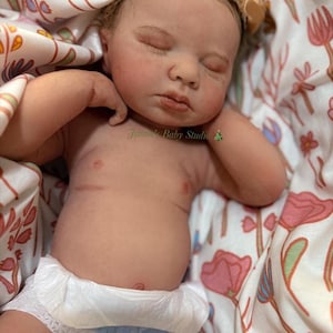 May include: A realistic baby doll with closed eyes, wearing a nappy, and a floral headband. The doll is lying on a floral patterned blanket. The text "Janna's Baby Studio" is visible on the image.