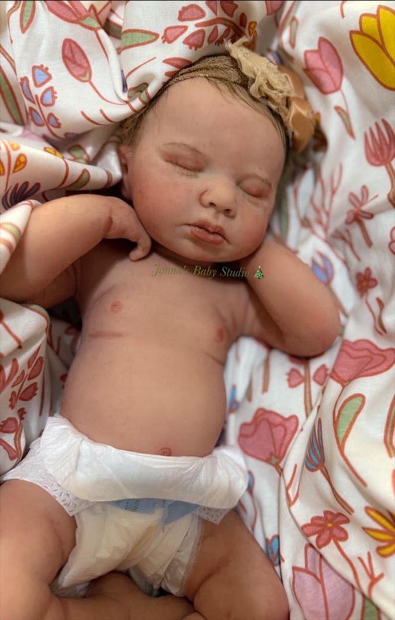 May include: A realistic baby doll with closed eyes, wearing a nappy, and a floral headband. The doll is lying on a floral patterned blanket. The text "Janna's Baby Studio" is visible on the image.