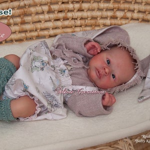 May include: A reborn baby doll named Kelsey Awake, dressed in a pink sweater, a white dress with a floral print, and green knitted socks. The doll is lying in a wicker basket with a white blanket. The doll has blue eyes and a realistic appearance. The image also includes a close-up of a real baby girl.