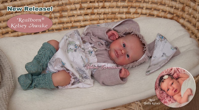 May include: A reborn baby doll named Kelsey Awake, dressed in a pink sweater, a white dress with a floral print, and green knitted socks. The doll is lying in a wicker basket with a white blanket. The doll has blue eyes and a realistic appearance. The image also includes a close-up of a real baby girl.