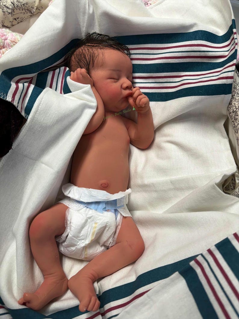 May include: A newborn baby doll with dark hair and rosy cheeks, wearing a diaper, lies swaddled in a white blanket with teal and burgundy stripes. The baby is resting with its eyes closed, and its hand is near its mouth.