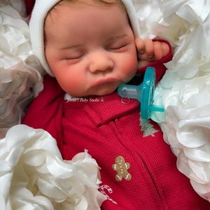 May include: A lifelike baby doll wearing a red onesie with a gingerbread man and Christmas tree applique. The doll has a white beanie and a blue pacifier. The doll is nestled in white fabric flowers. The text "Jamma's Baby Studio" is visible.
